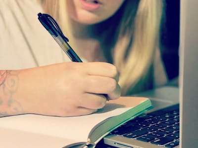 A person is writing in a notebook with a pen while using a laptop. The focus is on the hand and the writing action. A part of the person's face is visible, showcasing concentration.