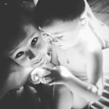 Two young children are seen in a close, intimate setting, with one child looking up with a soft expression and the other child focusing on something in their hands. The lighting is natural, casting gentle shadows.