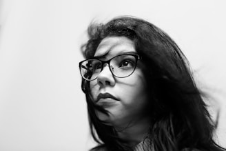A black and white portrait of a person with long dark hair wearing glasses, looking upwards with a thoughtful expression. The lighting highlights the texture of the hair and the clarity of the glasses, creating a contrast with the blurred white background.
