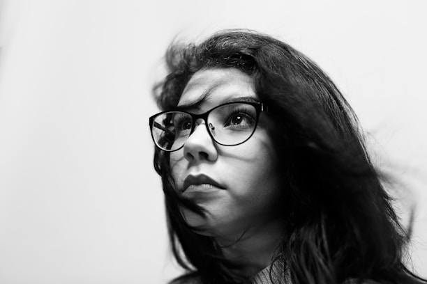 A black and white portrait of a person with long dark hair wearing glasses, looking upwards with a thoughtful expression. The lighting highlights the texture of the hair and the clarity of the glasses, creating a contrast with the blurred white background.