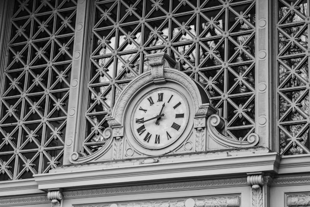 An ornate clock is set in a decorative framework with Roman numerals and intricate architectural details. The background features a pattern of geometric shapes, suggesting a wrought iron or lattice design.