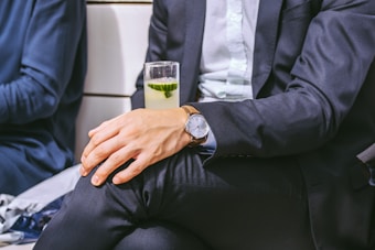 A person in a formal suit is sitting with legs crossed, holding a drink with a slice of cucumber in a tall glass. The person is wearing a watch on the left wrist.