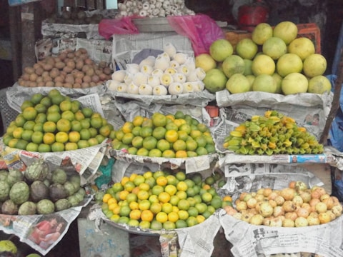 A vibrant display of various fruits arranged on a market stall. The fruits are neatly piled on top of newspaper sheets, including oranges, pomelos, sapodillas, mangoes, and custard apples. The arrangement forms a colorful and textured composition.