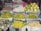 A vibrant display of various fruits arranged on a market stall. The fruits are neatly piled on top of newspaper sheets, including oranges, pomelos, sapodillas, mangoes, and custard apples. The arrangement forms a colorful and textured composition.