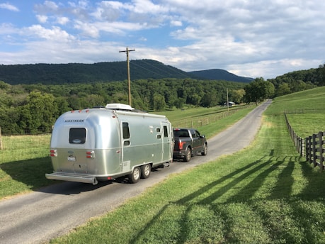 A scenic rural landscape with a silver Airstream trailer being towed by a dark-colored pickup truck on a narrow, paved country road. The road flanked by lush green fields and wooden fences leads towards distant rolling hills under a partly cloudy sky.