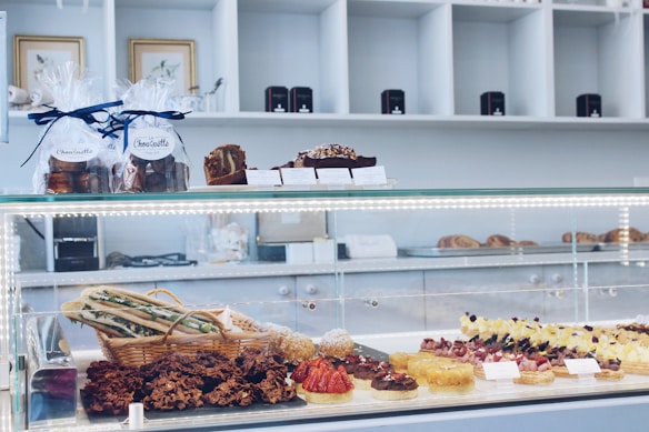 A display counter filled with a variety of pastries and baked goods, including tarts and other desserts. The upper shelves have packaged treats tied with ribbons, and the background shows a neatly organized, modern bakery setting.