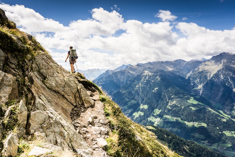 A hiker with a backpack and trekking poles is traversing a narrow mountain path. The path is rough and rocky, with patches of grass on either side. In the background, a panoramic view of a vast mountain range can be seen under a sky scattered with fluffy clouds.