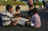 Four children are sitting on the ground playing a game of carrom on a board. The setting appears to be outdoors, possibly on a street, with a backdrop of some greenery and a stone wall. The children are focused on the game, and one of them is holding the striker.