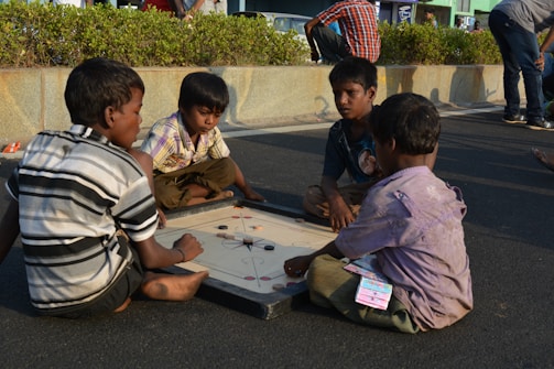 Four children are sitting on the ground playing a game of carrom on a board. The setting appears to be outdoors, possibly on a street, with a backdrop of some greenery and a stone wall. The children are focused on the game, and one of them is holding the striker.
