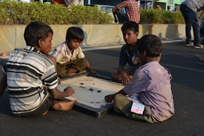 Four children are sitting on the ground playing a game of carrom on a board. The setting appears to be outdoors, possibly on a street, with a backdrop of some greenery and a stone wall. The children are focused on the game, and one of them is holding the striker.