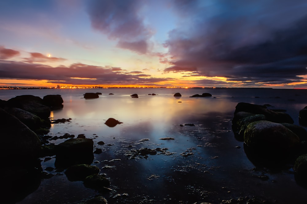 the sun is setting over the ocean with rocks in the foreground