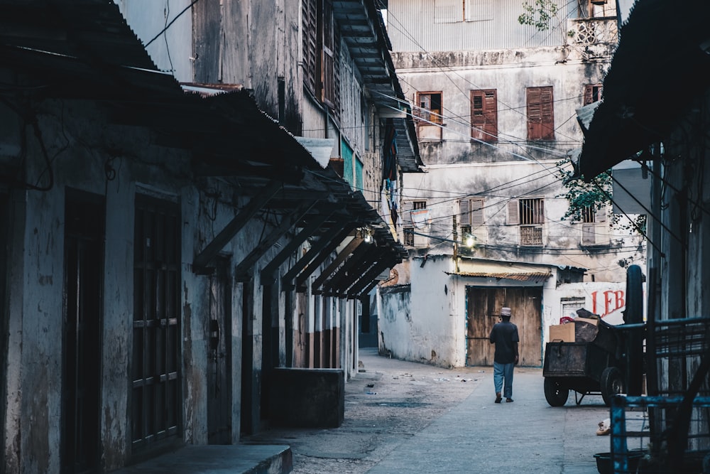 man walking near brown wooden cart between gray concrete buildings during daytime