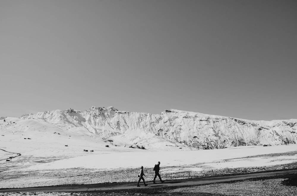 grayscale photography of two people walking near mountain covered snow