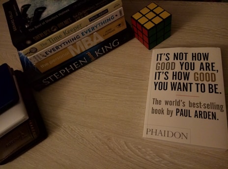 A collection of books is neatly arranged on a wooden surface. A Rubik's Cube is placed beside the stack. A book titled 'It's Not How Good You Are, It's How Good You Want to Be' by Paul Arden is prominently displayed on the right.