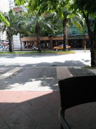 A street scene featuring several tall palm trees lining a sidewalk in front of a commercial building. The building has glass windows and a sign with visible text. A parked red car is seen in front of the building, while a motorcycle with a rider is passing by. The area is shaded by the trees, and there are seating arrangements with wicker chairs visible in the foreground.