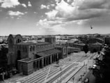 A historic stone church is set in an urban landscape. The church features arched windows and detailed architectural elements. A cobbled street and sidewalk lead away from the church, with some pedestrians and parked cars visible. The cityscape extends into the background, showing a mix of old and modern buildings. A bird is in mid-flight against a cloudy sky.