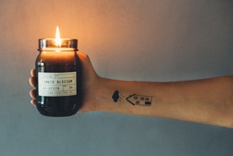 A hand holding a lit candle jar labeled 'Tomato Blossom.' The arm has a tattoo featuring a house and a cloud with rain on it. The background is softly blurred, with a warm glow cast by the candlelight.