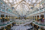 A spacious and elegant shopping mall features intricate architectural designs with a large arched glass ceiling. The interior includes multiple levels of shop facades with decorative railings and ambient spherical lamps. Lush green plants add a touch of nature to the setting, while natural light floods the area.