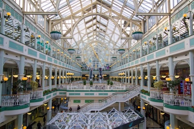 A spacious and elegant shopping mall features intricate architectural designs with a large arched glass ceiling. The interior includes multiple levels of shop facades with decorative railings and ambient spherical lamps. Lush green plants add a touch of nature to the setting, while natural light floods the area.
