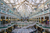 A spacious and elegant shopping mall features intricate architectural designs with a large arched glass ceiling. The interior includes multiple levels of shop facades with decorative railings and ambient spherical lamps. Lush green plants add a touch of nature to the setting, while natural light floods the area.