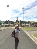 A woman with long hair is standing on a paved pathway at a theme park entrance, wearing a white cap, a light gray t-shirt, black pants, and carrying a colorful backpack. The background features a row of ornate buildings with gabled roofs and a Ferris wheel behind them. Two tall flags are prominently displayed on flagpoles: one is the flag of the Philippines, and the other is a park flag.