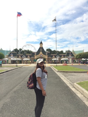 A woman with long hair is standing on a paved pathway at a theme park entrance, wearing a white cap, a light gray t-shirt, black pants, and carrying a colorful backpack. The background features a row of ornate buildings with gabled roofs and a Ferris wheel behind them. Two tall flags are prominently displayed on flagpoles: one is the flag of the Philippines, and the other is a park flag.