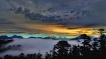 The image features a mountainous landscape with a dramatic, colorful sunset sky transitioning from warm hues of orange and yellow to cooler shades of blue and purple. Layers of clouds hang low, partially obscuring the peaks, while silhouettes of trees add depth to the foreground.