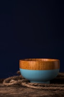 A wooden bowl with a blue painted exterior resting on a wooden surface with a piece of rope lying around it. The background is dark, creating a strong contrast against the bowl.
