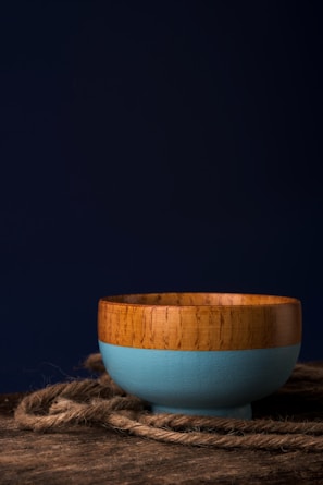 A wooden bowl with a blue painted exterior resting on a wooden surface with a piece of rope lying around it. The background is dark, creating a strong contrast against the bowl.