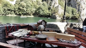 A scenic riverside view featuring a variety of items on wooden tables, including textiles, a globe, wooden boxes, and an ornate clock. The background showcases lush greenery and rocky cliffs under clear skies, with a river flowing gently. A lone lamp post stands near the river's edge while a person is partially visible on the right side.