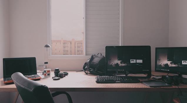 A workspace featuring multiple computer monitors, a laptop, and various photography equipment is placed on a wooden desk. The scene includes a keyboard, a backpack, headphones, and a window with a view of buildings under an overcast sky.