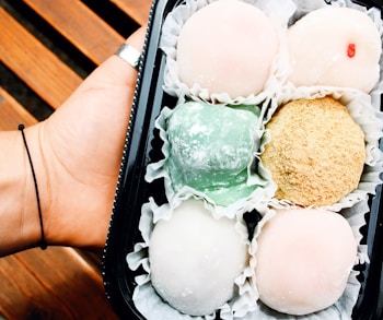 A hand holding a black tray containing six pieces of colorful mochi, each resting in a crinkled paper cup. The mochi are in pastel shades of pink, green, beige, and white. The wooden background adds a natural element to the presentation.