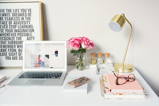 A neatly arranged desk features a laptop displaying a website, a vase of pink roses, a brass desk lamp, and various office supplies. There's a framed motivational quote on the wall behind and notebooks with glasses and a smartphone on the desk.