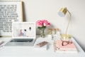 A neatly arranged desk features a laptop displaying a website, a vase of pink roses, a brass desk lamp, and various office supplies. There's a framed motivational quote on the wall behind and notebooks with glasses and a smartphone on the desk.