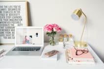 A neatly arranged desk features a laptop displaying a website, a vase of pink roses, a brass desk lamp, and various office supplies. There's a framed motivational quote on the wall behind and notebooks with glasses and a smartphone on the desk.