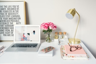 A neatly arranged desk features a laptop displaying a website, a vase of pink roses, a brass desk lamp, and various office supplies. There's a framed motivational quote on the wall behind and notebooks with glasses and a smartphone on the desk.