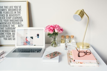 A neatly arranged desk features a laptop displaying a website, a vase of pink roses, a brass desk lamp, and various office supplies. There's a framed motivational quote on the wall behind and notebooks with glasses and a smartphone on the desk.