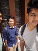 Three young men are standing indoors with a rocky background wall. They are casually dressed and smiling, suggesting a friendly environment. Two of them are wearing backpacks, and there is a contrast between dark and light clothing.