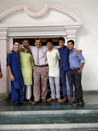 A group of six men are standing together on a set of steps in front of a building with an arched entrance. They are smiling and have their arms around each other, suggesting a friendly or celebratory moment. The men are dressed in a variety of traditional and modern clothing, including a tie, kurta, and formal shirts.