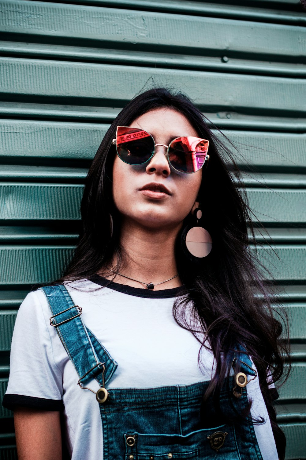 a woman wearing sunglasses and overalls standing in front of a green wall