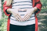A person wearing a red jacket and a striped shirt is forming a heart shape with their hands over their abdomen, standing outside in a natural setting.