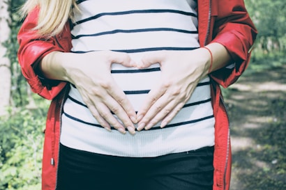 A person wearing a red jacket and a striped shirt is forming a heart shape with their hands over their abdomen, standing outside in a natural setting.