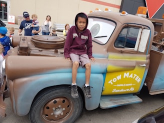 A child wearing a maroon hoodie and shorts is sitting on a large, character-themed vehicle resembling a tow truck from an animated movie. The truck has a friendly cartoon face on its windshield and is painted in shades of brown and blue with the words 'Tow Mater' on the side. People, including children, are gathered around, and some are observing and smiling.