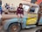 A child wearing a maroon hoodie and shorts is sitting on a large, character-themed vehicle resembling a tow truck from an animated movie. The truck has a friendly cartoon face on its windshield and is painted in shades of brown and blue with the words 'Tow Mater' on the side. People, including children, are gathered around, and some are observing and smiling.