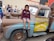 A child wearing a maroon hoodie and shorts is sitting on a large, character-themed vehicle resembling a tow truck from an animated movie. The truck has a friendly cartoon face on its windshield and is painted in shades of brown and blue with the words 'Tow Mater' on the side. People, including children, are gathered around, and some are observing and smiling.