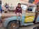 A child wearing a maroon hoodie and shorts is sitting on a large, character-themed vehicle resembling a tow truck from an animated movie. The truck has a friendly cartoon face on its windshield and is painted in shades of brown and blue with the words 'Tow Mater' on the side. People, including children, are gathered around, and some are observing and smiling.