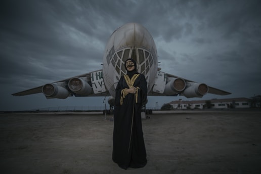 A person dressed in black traditional clothing with gold accents stands in front of an old, possibly abandoned airplane. The airplane has worn graffiti on it and looks weathered. The sky is overcast, contributing to a gloomy atmosphere. There are buildings in the background, suggesting an airfield or base.