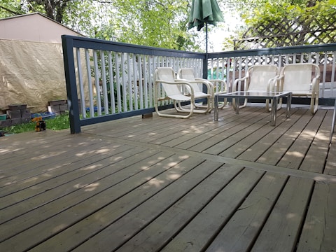 A wooden deck with several white patio chairs and a glass-top table. The area is surrounded by a railing, and there is a green umbrella providing shade. The background includes trees and a shed covered in a tarp.
