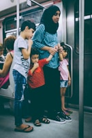 A woman wearing a headscarf stands in a train carriage holding hands with three children. One of the children is standing close to her in a red and black striped shirt, while another holds onto her other hand. The environment suggests a crowded public transport setting.