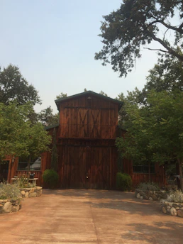 A rustic wooden barn surrounded by lush greenery and trees. The structure has large double doors and a prominent triangular roof. There are neatly arranged stone planters with shrubs on either side of the entrance. The setting appears calm and serene, situated on a paved area.